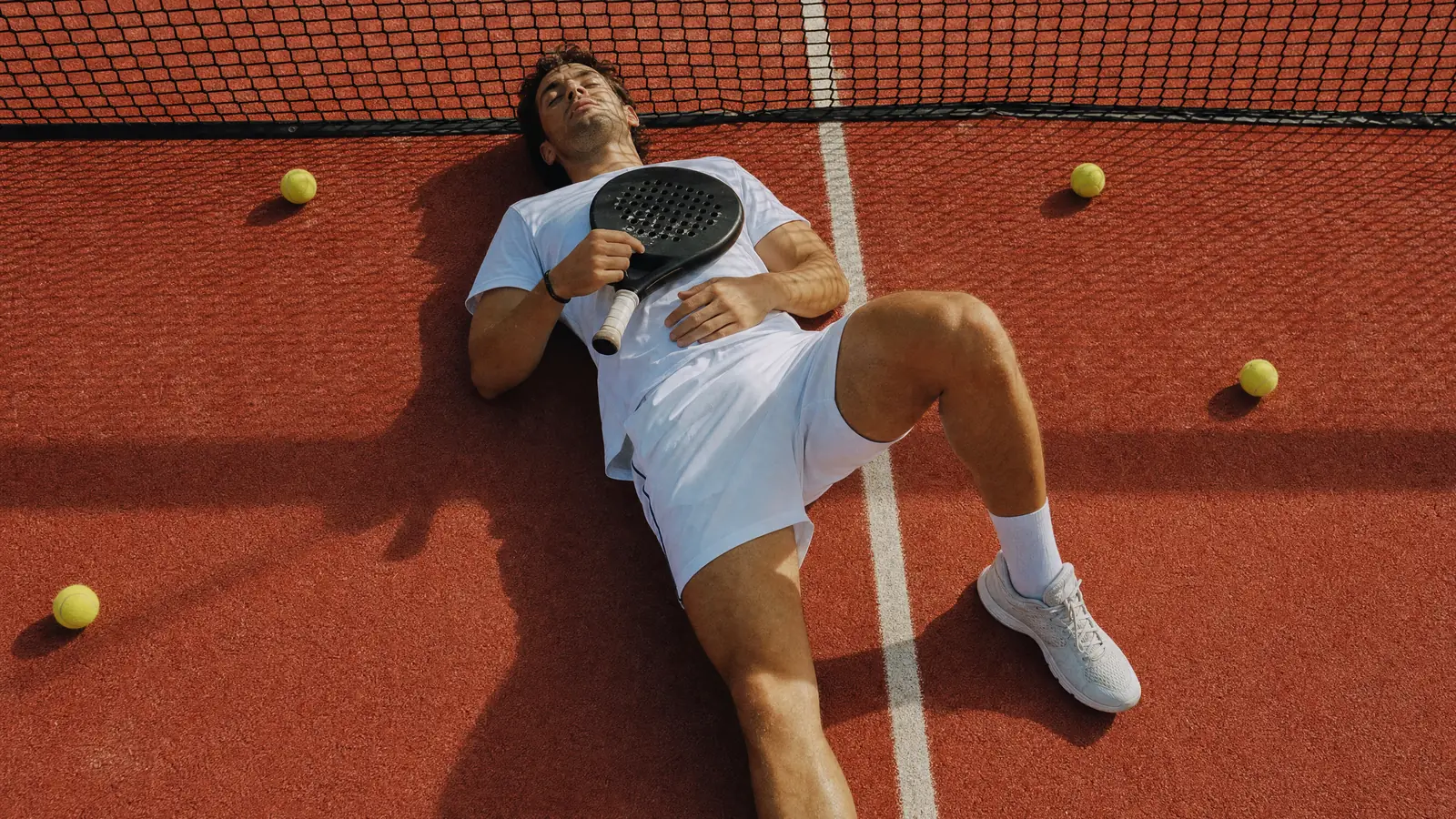 A padel player lying on his back on a red court under the net, racket on his chest, balls scattered around him