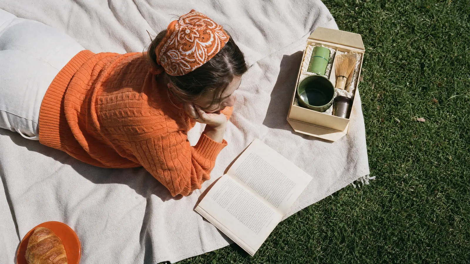 Seen from above, a woman in an orange bandana reading on a picnic blanket next to a boxed tea set and a croissant on an orange plate