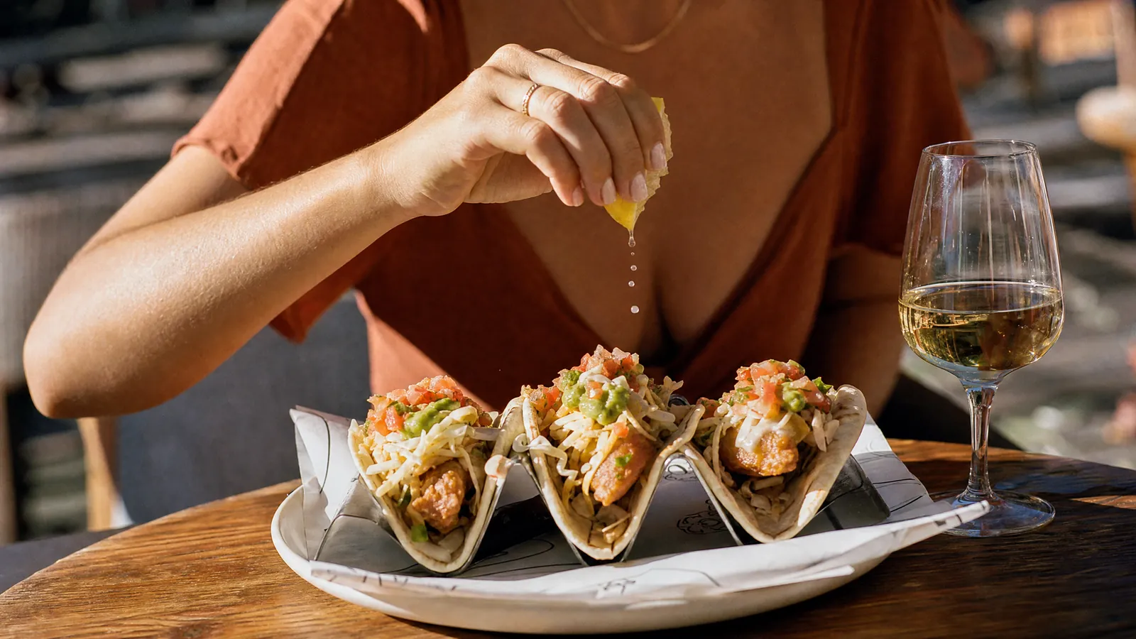 A hand squeezing a lemon over three fish tacos at a sunlit restaurant table, a glass of white wine beside the plate