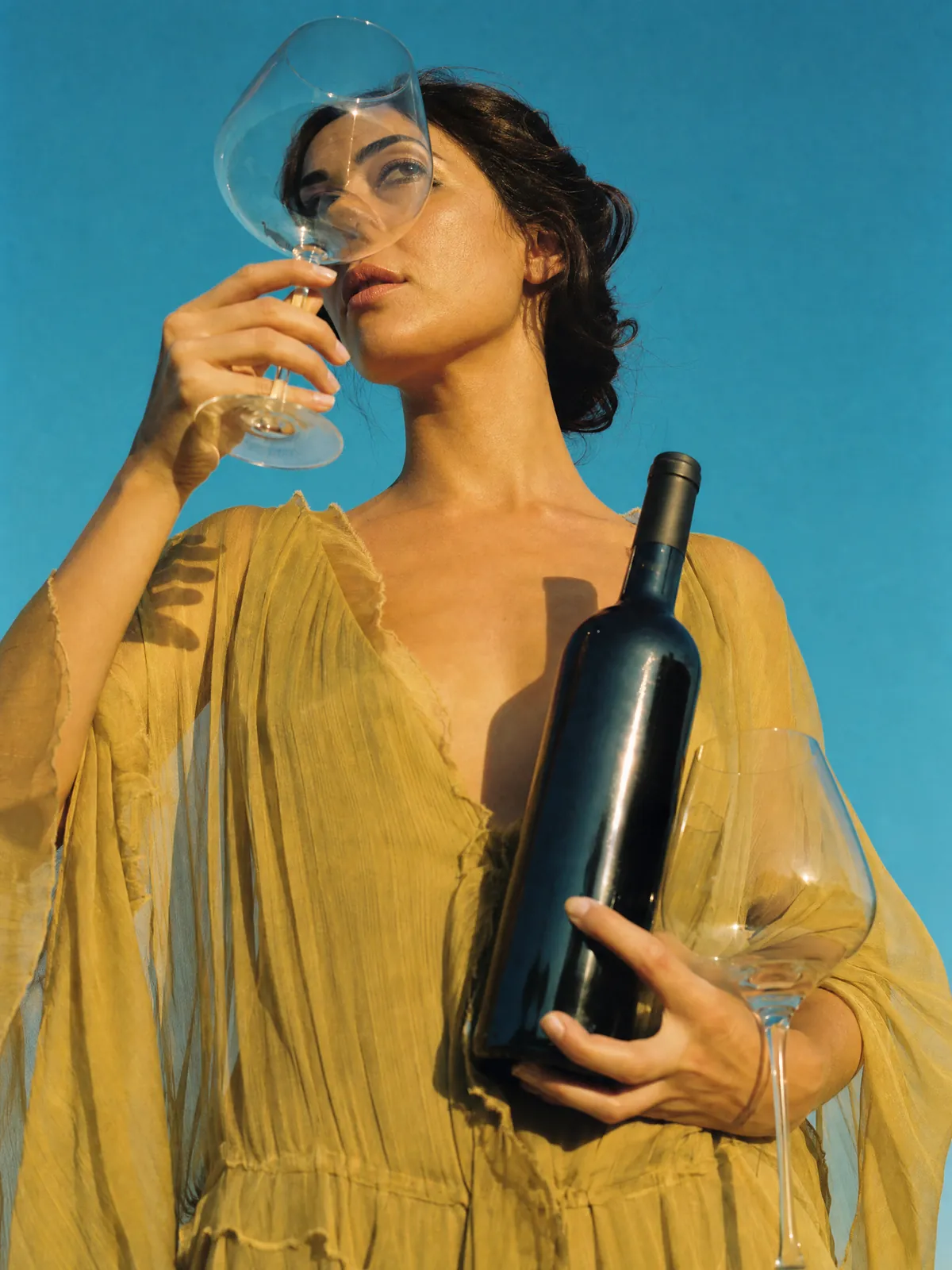 A woman in a sheer yellow dress against a clear sky, holding a bottle of red wine and two glasses, her face seen through one of them