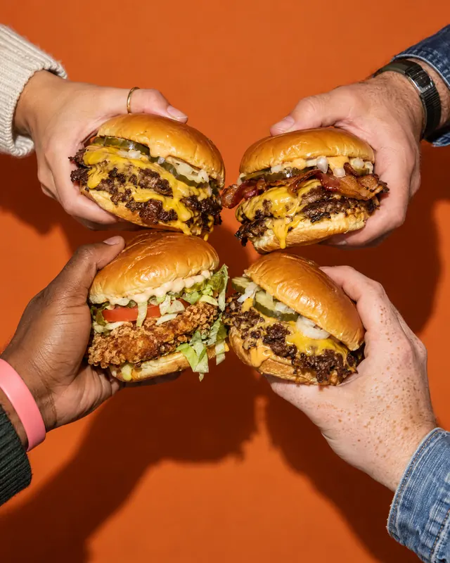 Four hands bring four smash burgers together on orange paper